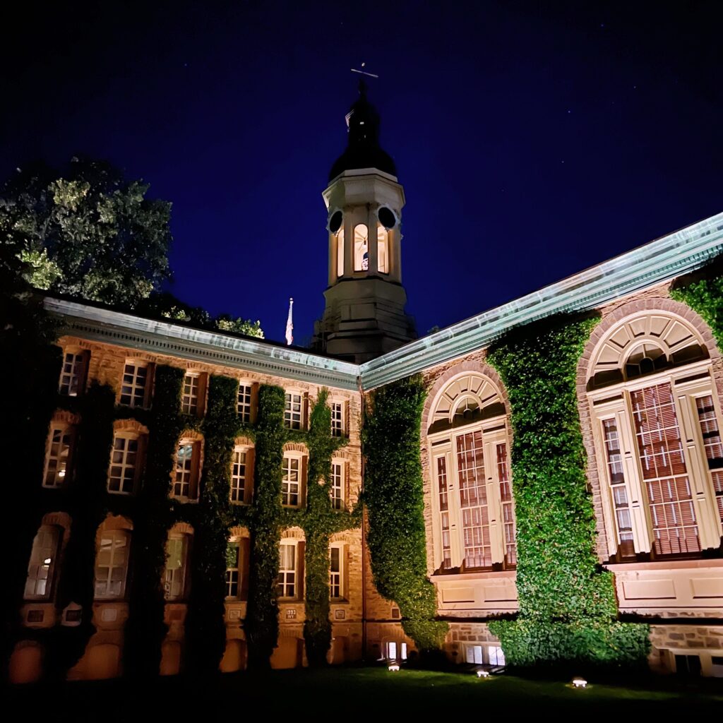 Photo depicts grand Princeton building at night time, with ivy climbing up brick that appears reddish in the lighting.