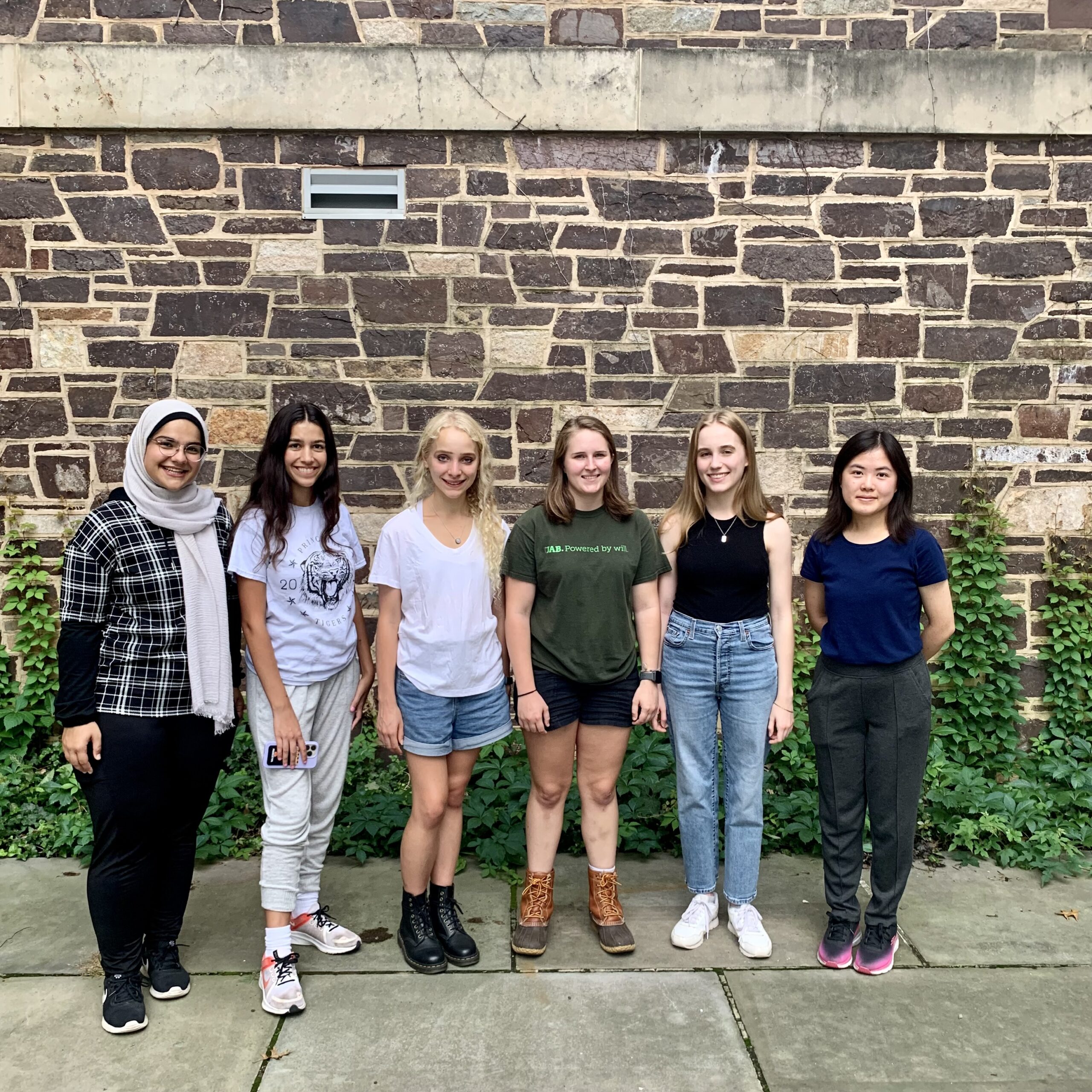 Image depicts six women (PCUR 22-23) in front of a gray brick building with bright green ivy