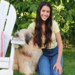 Image depicts young woman with long brown hair kneeling on the grass with her arm around a dog.
