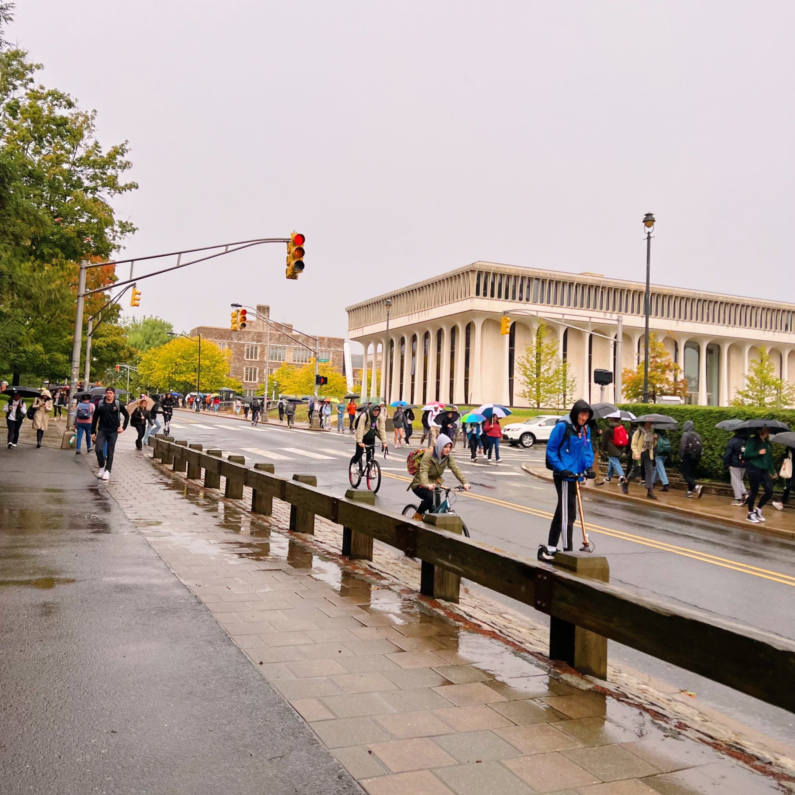 Photograph of Princeton University in the rain, with many students walking, biking, and scootering to class holding umbrellas or wearing rain jackets