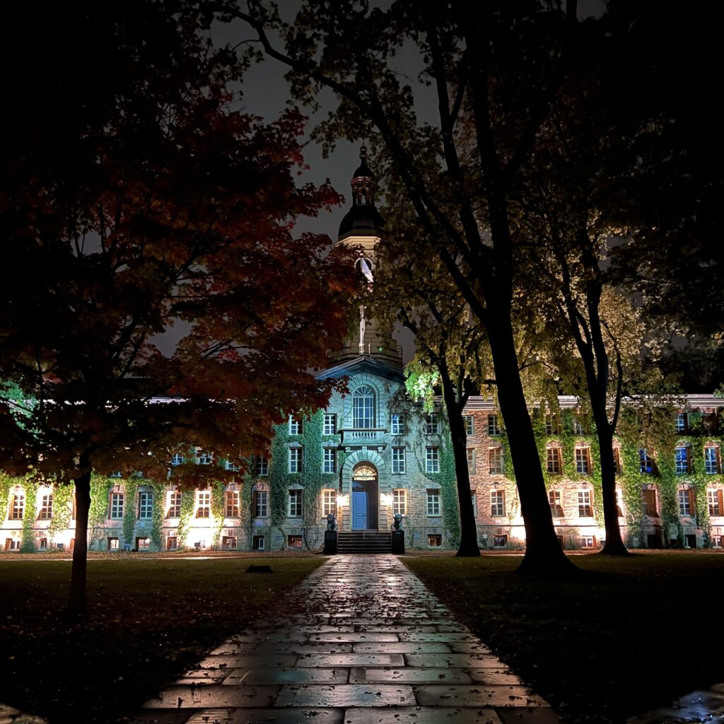 Photograph of Nassau Hall at night. Blueish-Green light is cast over the center of the building. The brick pathway leading up is wet and covered in fall leaves.