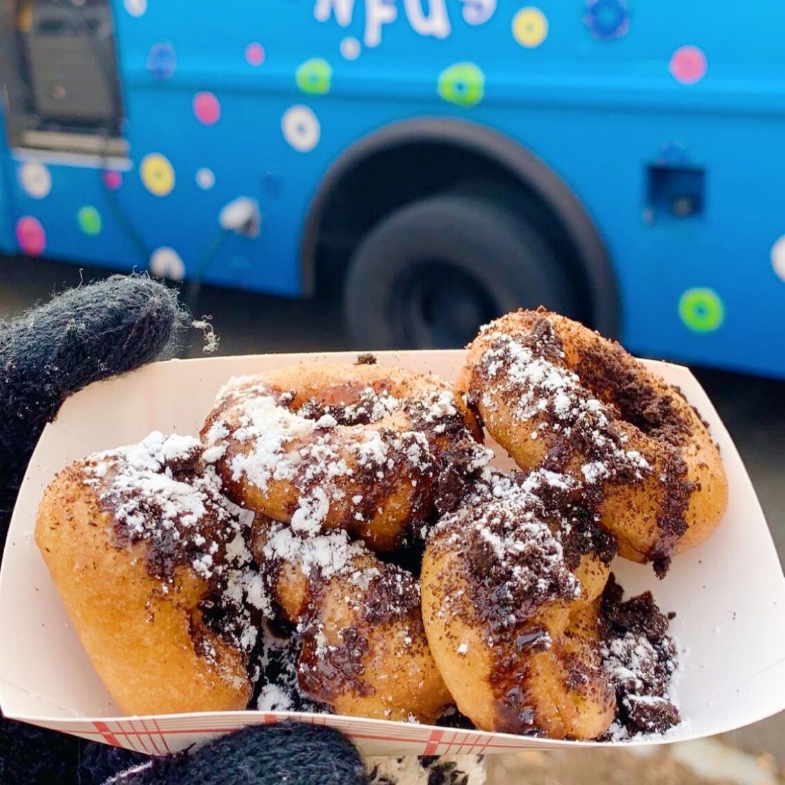 Photo of hand holding a takeout container with mini donuts covered in chocolate and powdered sugar. A blue food truck is in the background.