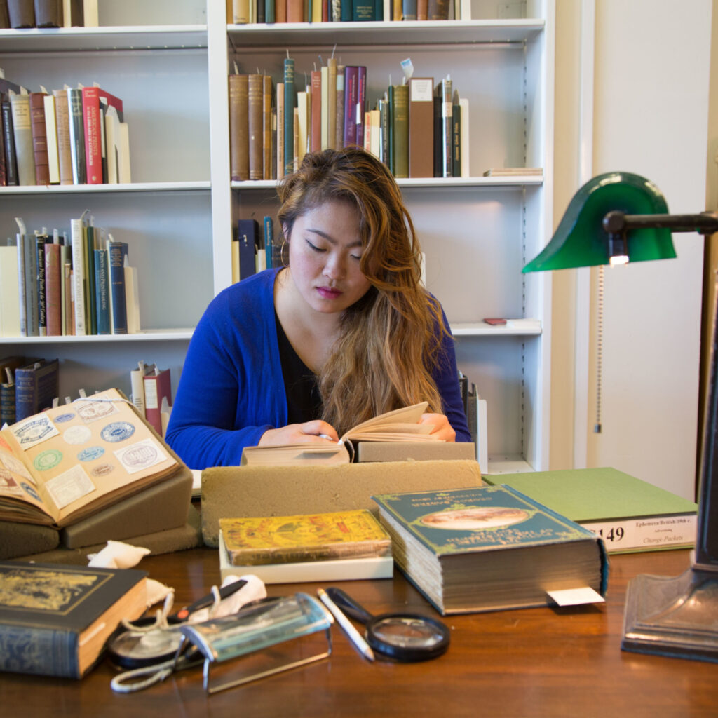 Image depicts student reading a book at a desk covered with texts and research tools, sitting in front of library shelves.