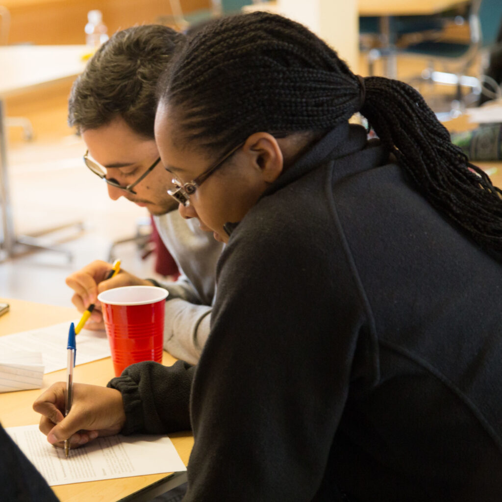 Two students sit at a table with their pen hovering over paper, working on their senior theses.