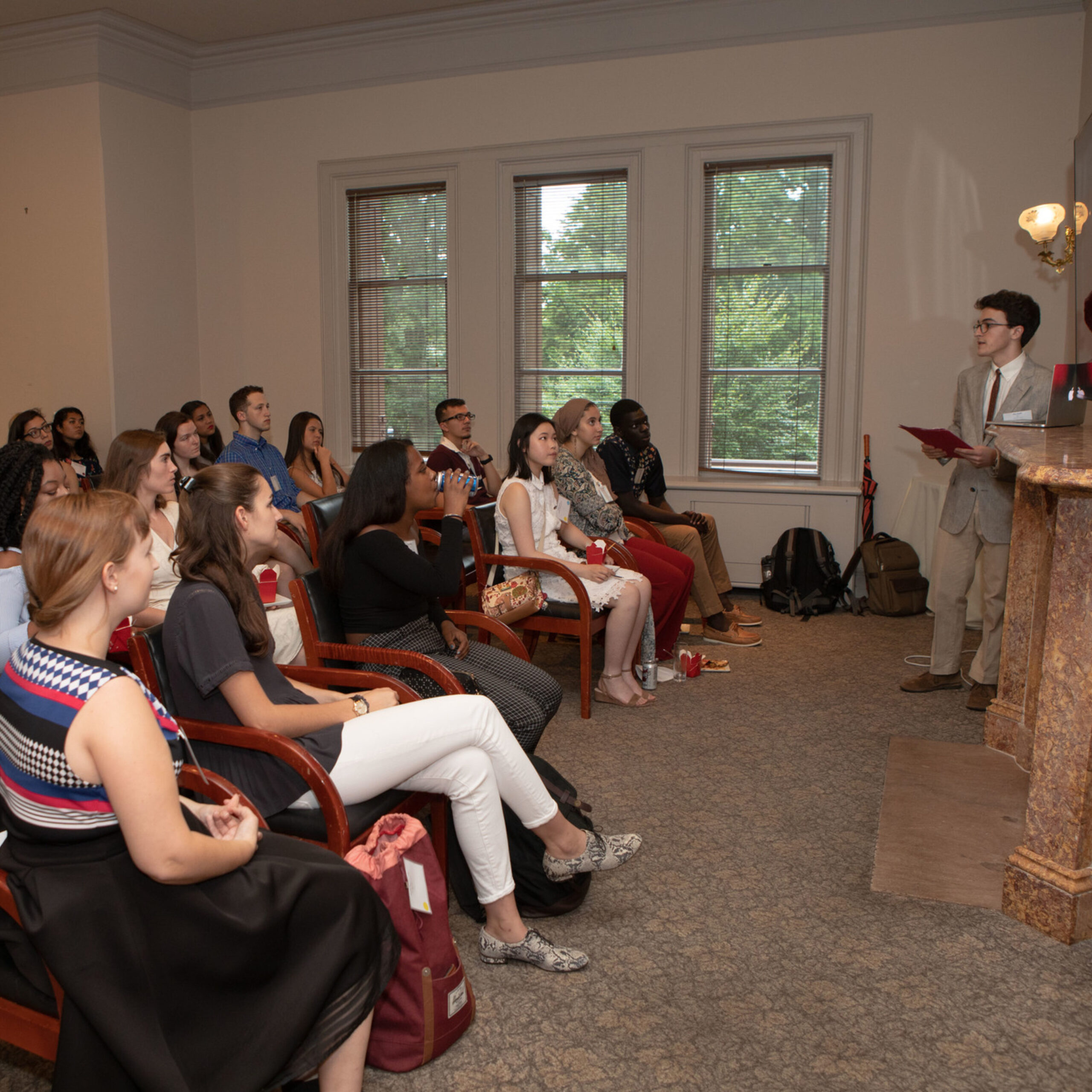 Students sitting in chairs face a man in a grey suit standing and speaking to them 