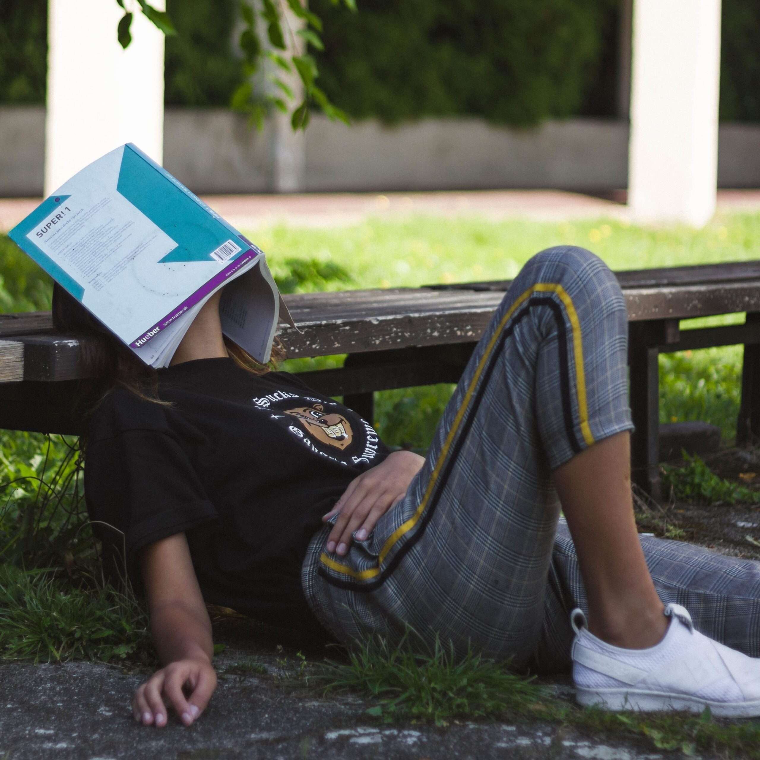 Photo of girl sitting on grass, leaning against bench, face covered by open textbook. 