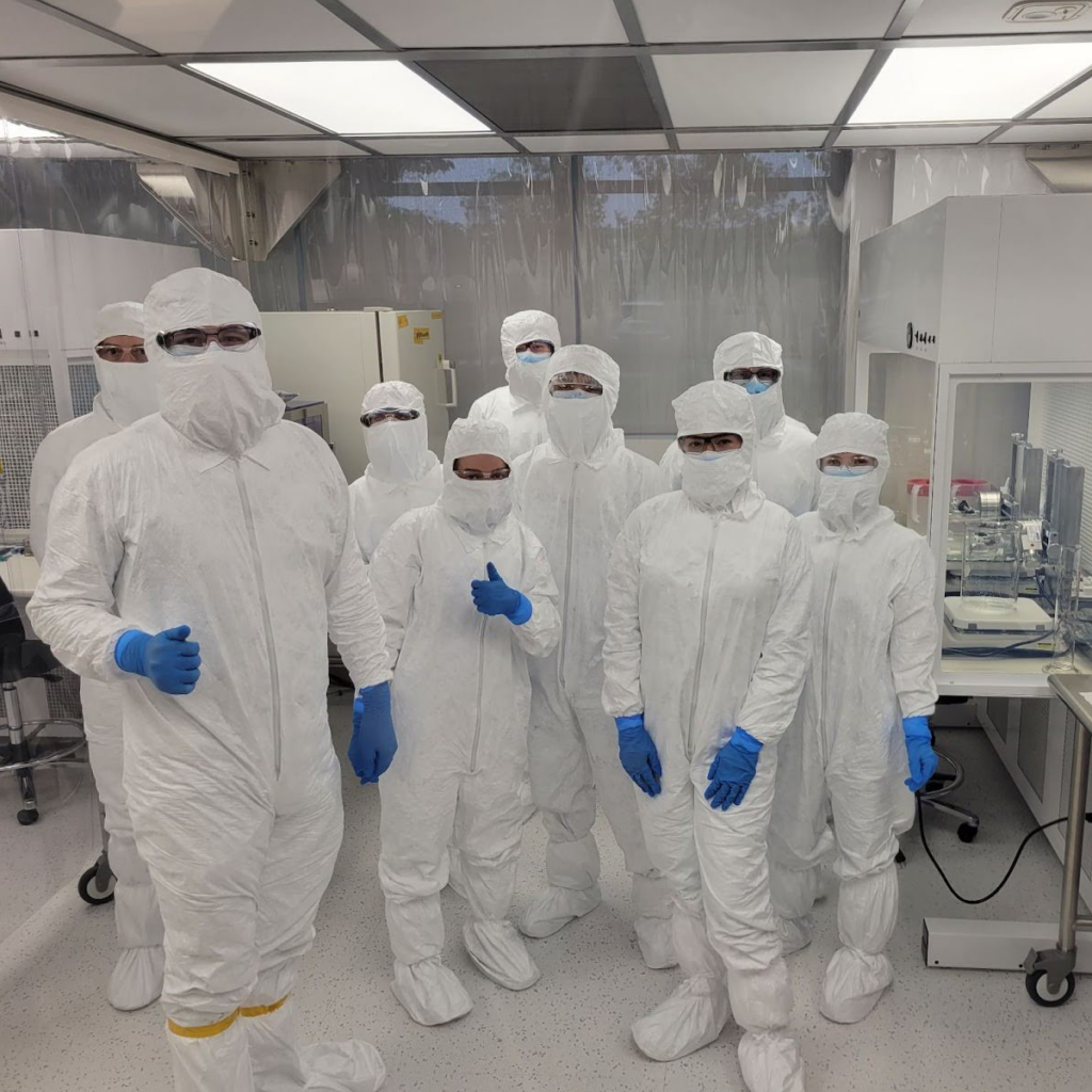 Eight students and a lab engineer are wearing white coverall suits while working in a cleanroom.