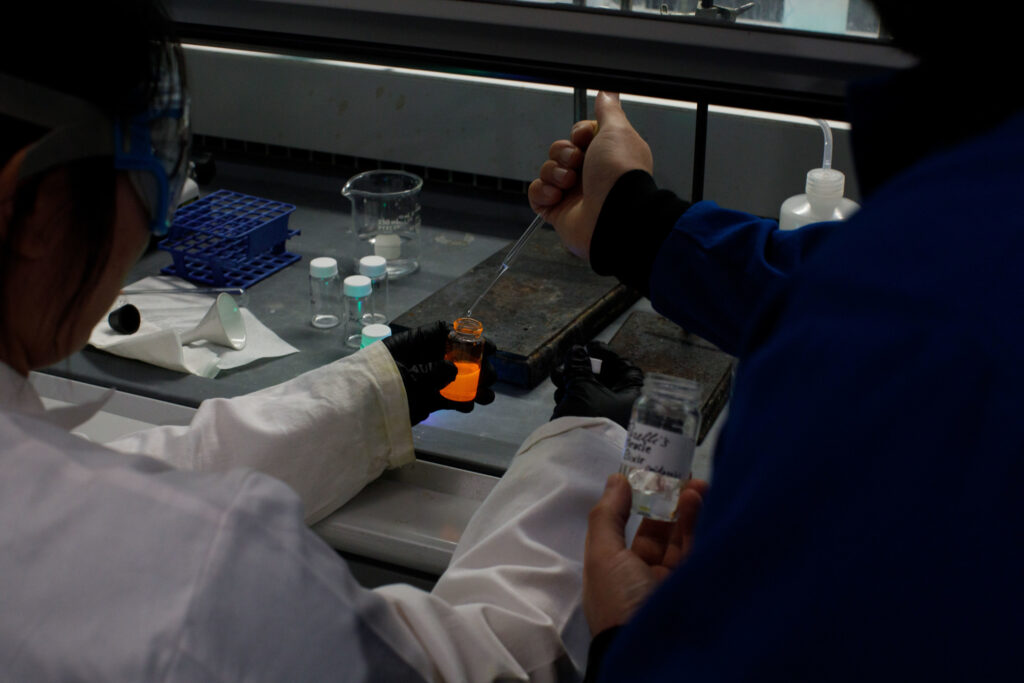 This image shows a lab technician pipetting a clear solution into an another orange-colored solution inside a fume hood in a lab