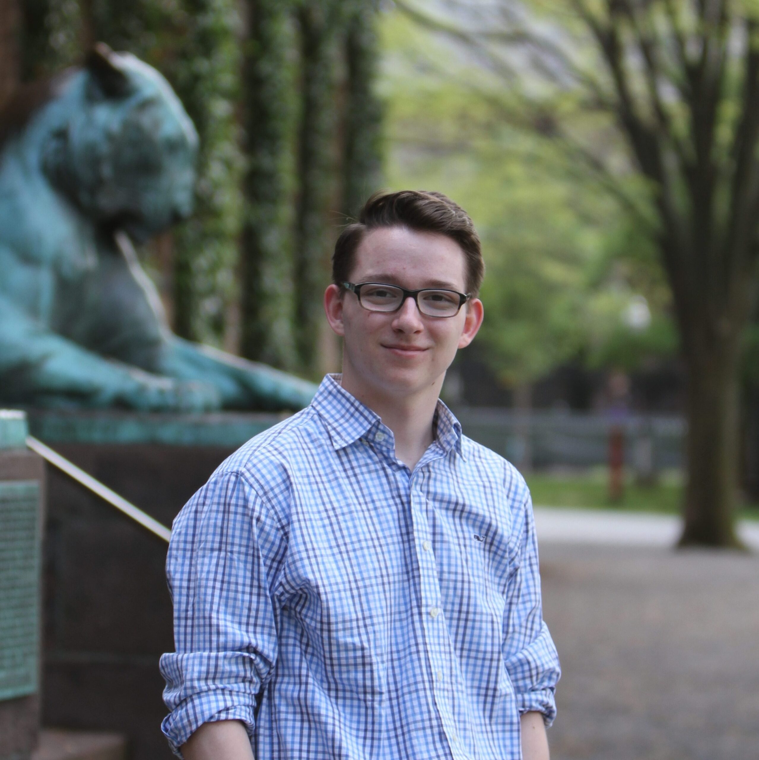 Portrait of a young man with short brown hair, a blue checkered dress shirt, smiling warmly at the camera in front of Nassau Hall.