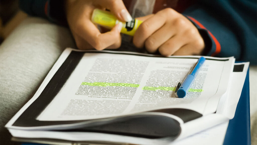 A printed article with yellow highlighting under a blue pen with the hands of the person reading and annotating.