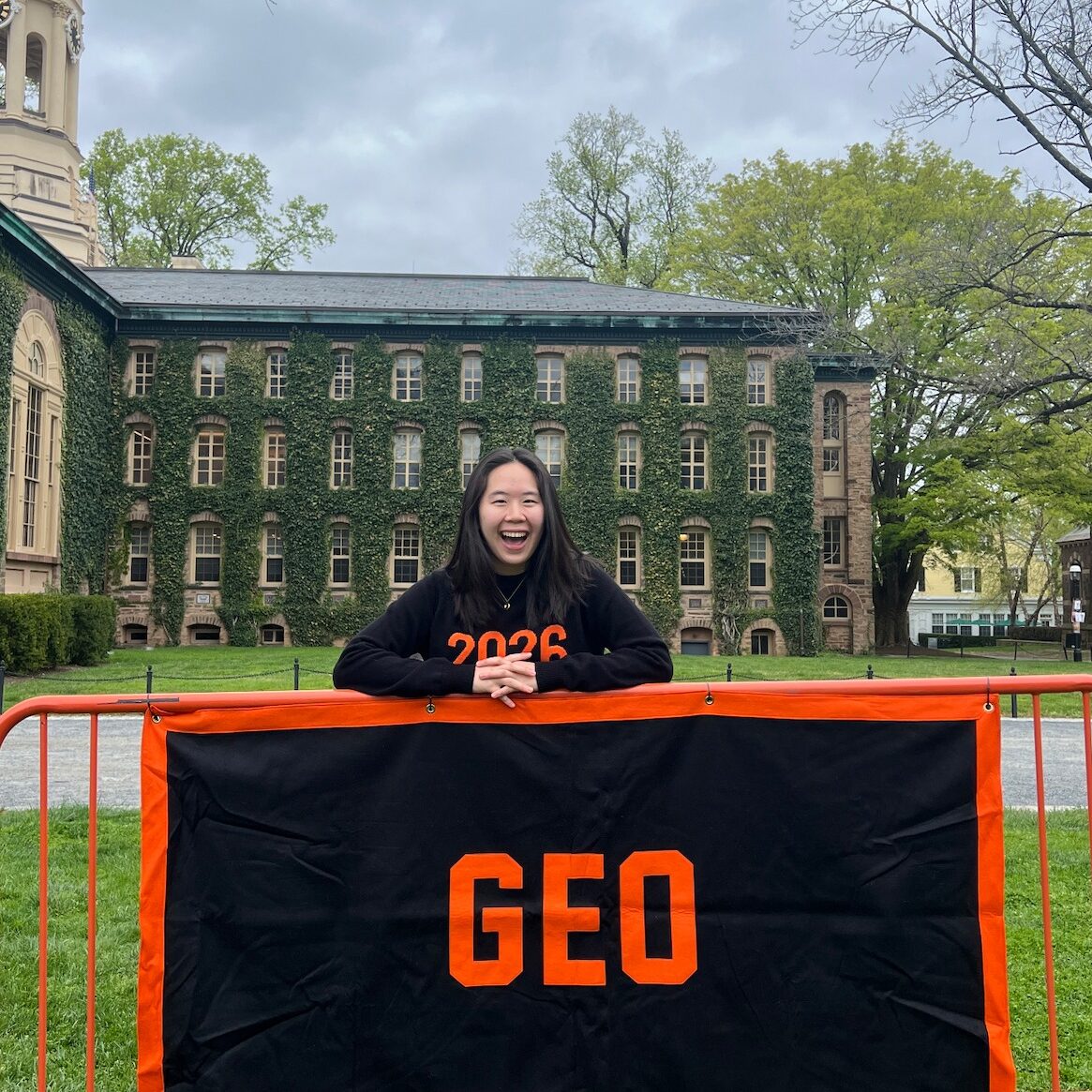 Picture features Rebecca wearing 2026 sweater posing with large sign that say "GEO" on canon green behind nassau hall.