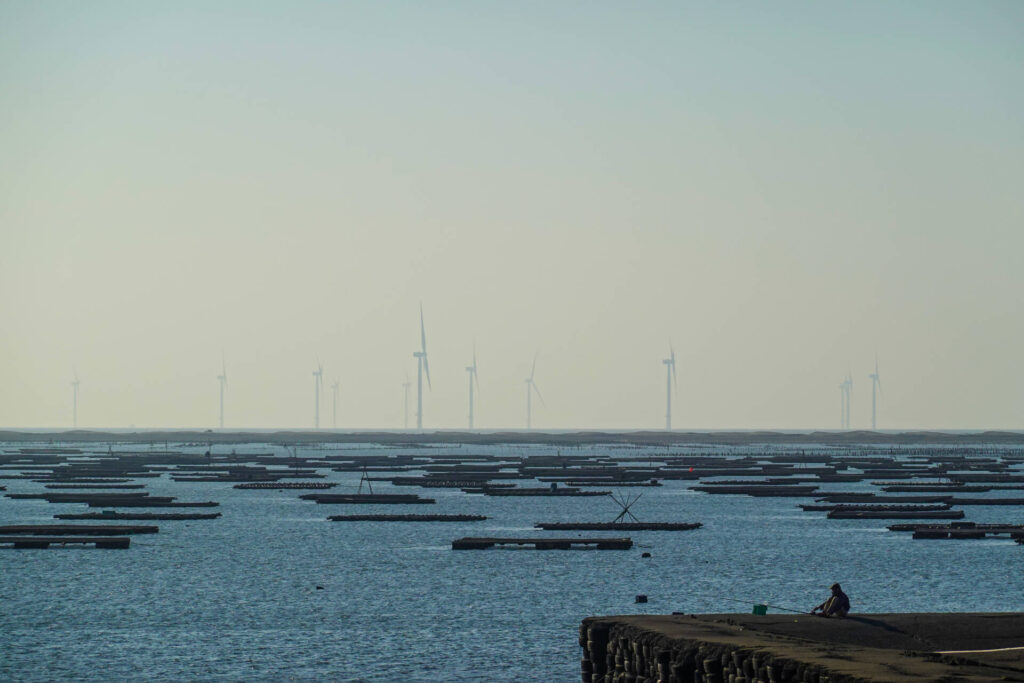Local fishermen sitting near fishing rod on shore, looking out into a horizon of oyster aquaculture and an offshore wind farm